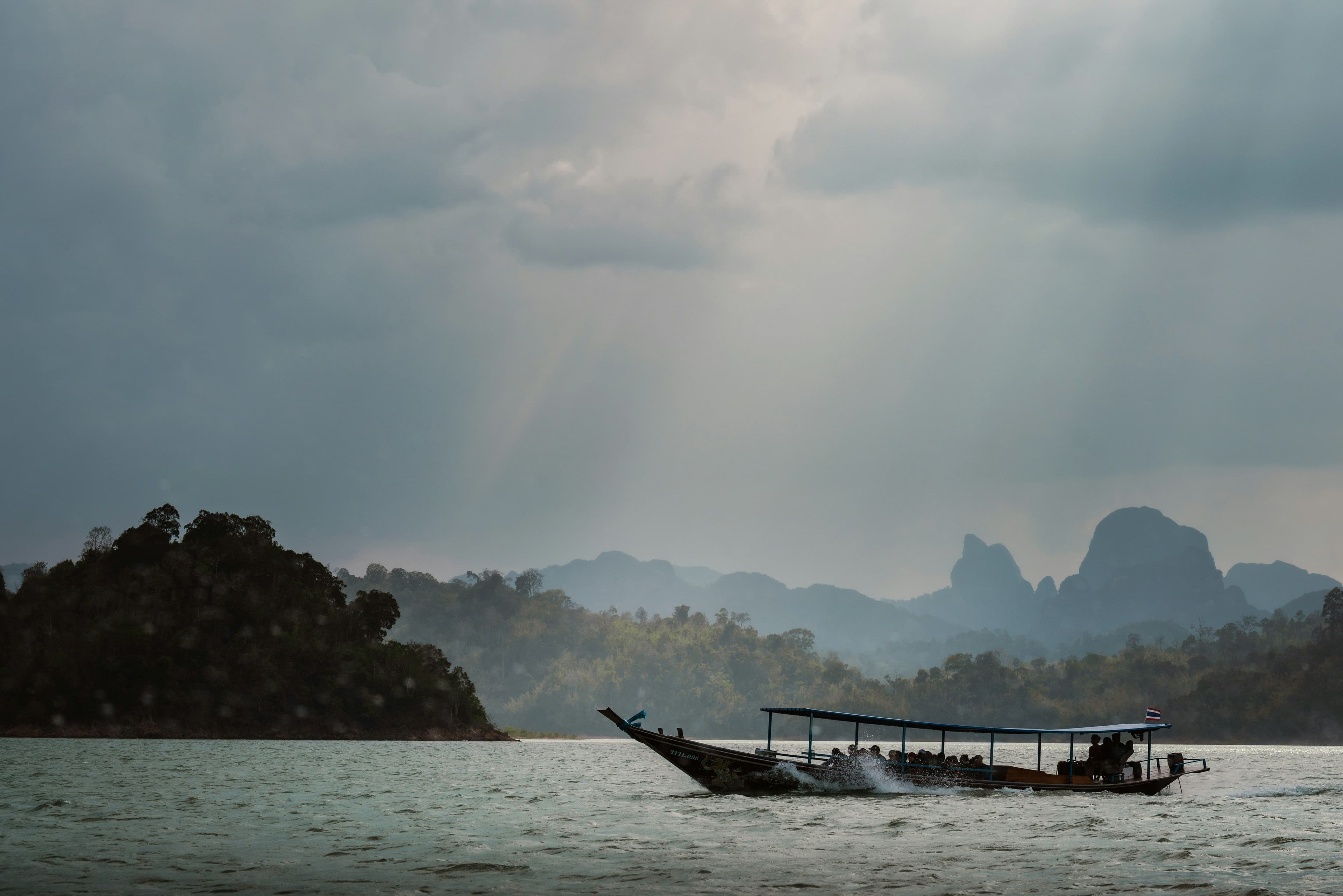 Longtail boat crossing a calm lake in Thailand, surrounded by lush forested islands and misty limestone mountains beneath dramatic storm clouds with sunlight streaming through.