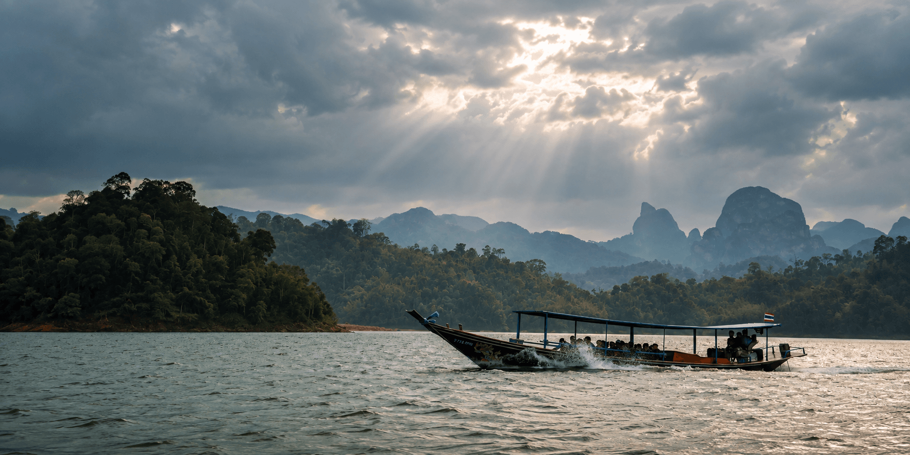 Longtail boat crossing a calm lake in Thailand, surrounded by lush forested islands and misty limestone mountains beneath dramatic storm clouds with sunlight streaming through.